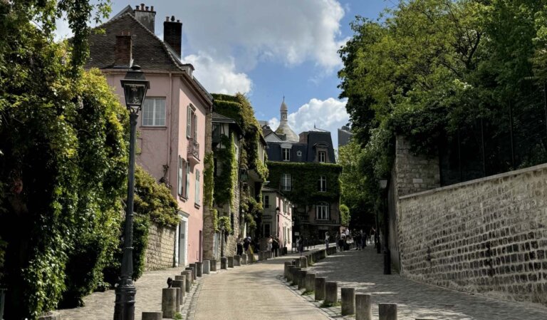 A winding street in Montmartre, Paris, with a stone wall on the right and the famous pink house covered in ivy on the left